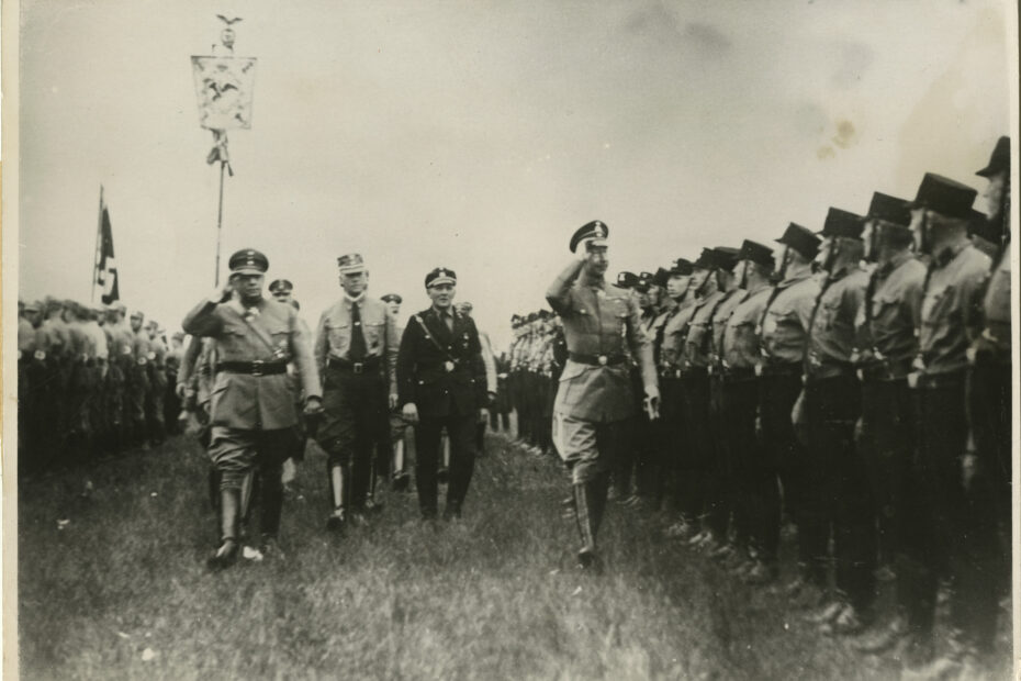 Auf der rechten und linken Seite stramm stehende Reihen von uniformierten Soldaten. Im Hintergrund ist eine Fahne mit Hakenkreuz zu sehen. In der Mitte Zug von Offizieren, die die Soldaten grüßen. Weiter hinten wird ein Wappen getragen.