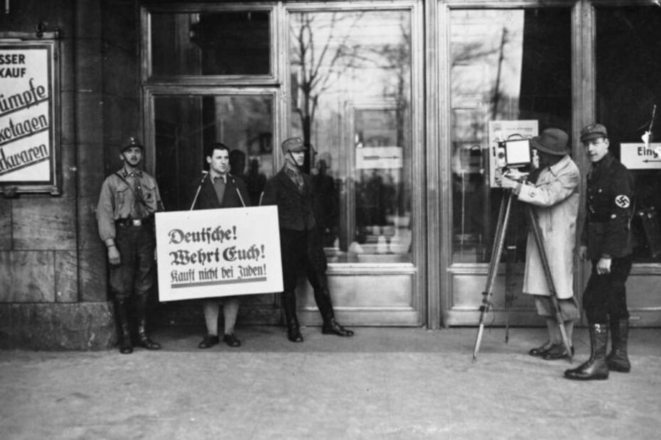 Bild aus 1933, das die Boykottaktion der Nazis gegen jüdische Geschäfte zeigt. SA- und SS-Leute vor Kaufhaus Wertheim, Mann mit Schild um den Hals "Deutsche! Wehrt Euch! Kauft nicht bei Juden!", rechts Mann mit Filmkamera filmend.