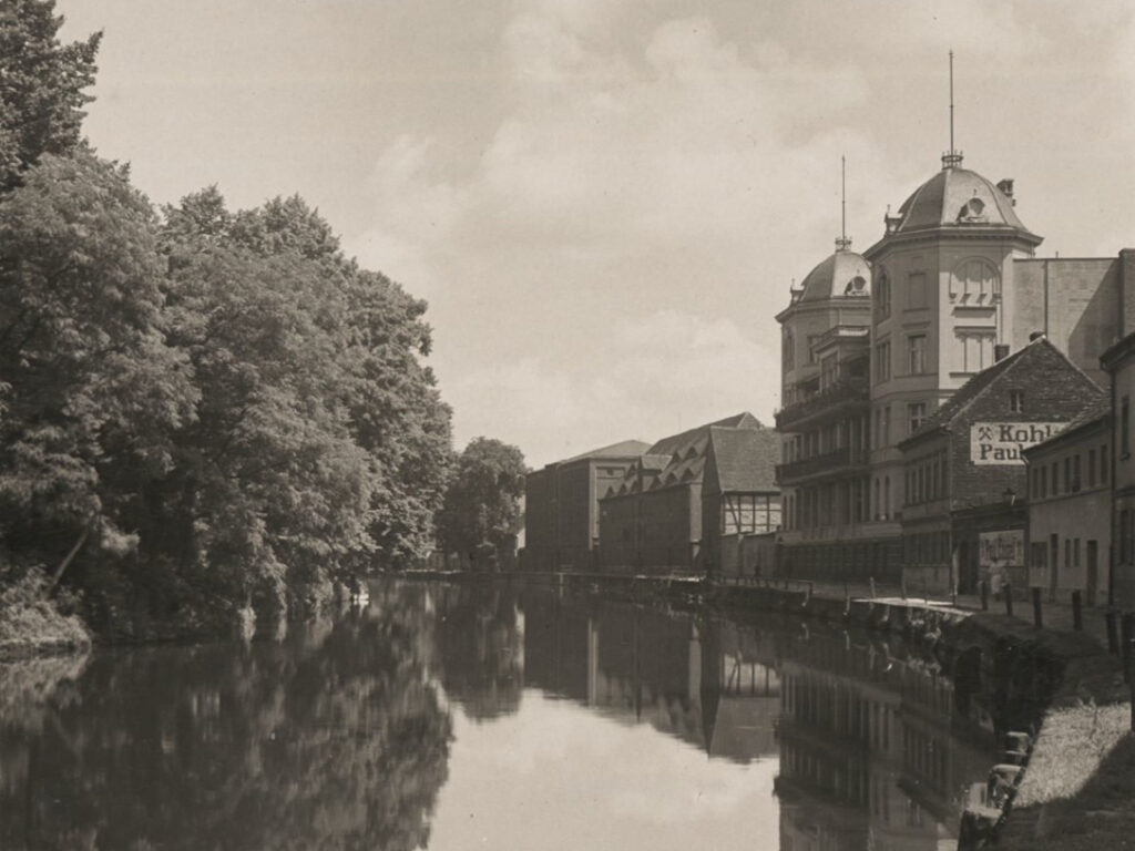 Bild zeigt einen Teil der Havel. Links von dem Fluss sind große Bäume und Gebüsch zu sehen. Rechts mehrere Häuser am Uferrand.