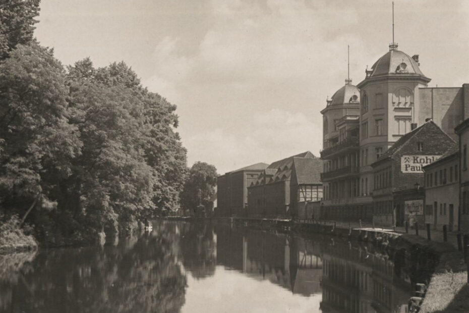 Bild zeigt einen Teil der Havel. Links von dem Fluss sind große Bäume und Gebüsch zu sehen. Rechts mehrere Häuser am Uferrand.