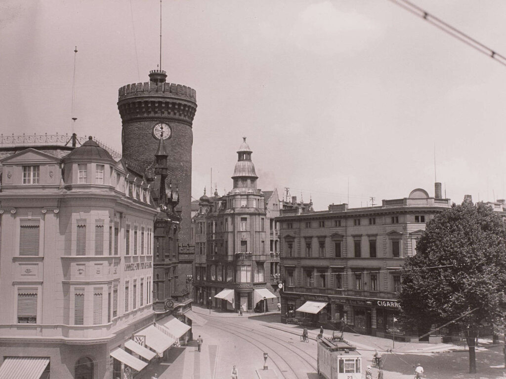 Bild aus 1935. Zeigt eine Einkaufsstraße von oben. Unten sieht man eine Straßenbahn durch die Straße fahren. Links Häuser und dahinter ein großer Turm.