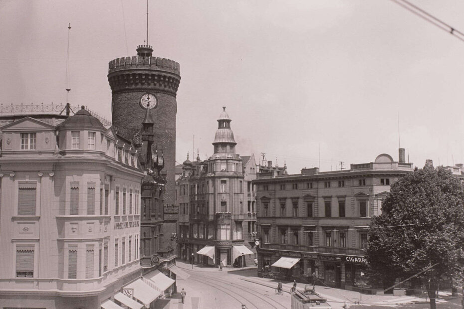 Bild aus 1935. Zeigt eine Einkaufsstraße von oben. Unten sieht man eine Straßenbahn durch die Straße fahren. Links Häuser und dahinter ein großer Turm.
