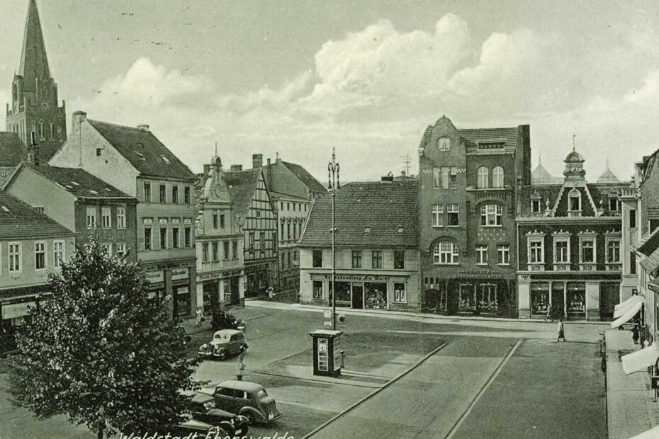 Bild, auf dem geschrieben steht "Waldstadt Eberswalde. Marktplatz mit Maria-Magdalenen-Kirche". In der Mitte des Bildes eine großer Platz mit Parkplätzen und einigen Autos. Der Platz wird von drei Seiten von Häuserreihen eingeschlossen.