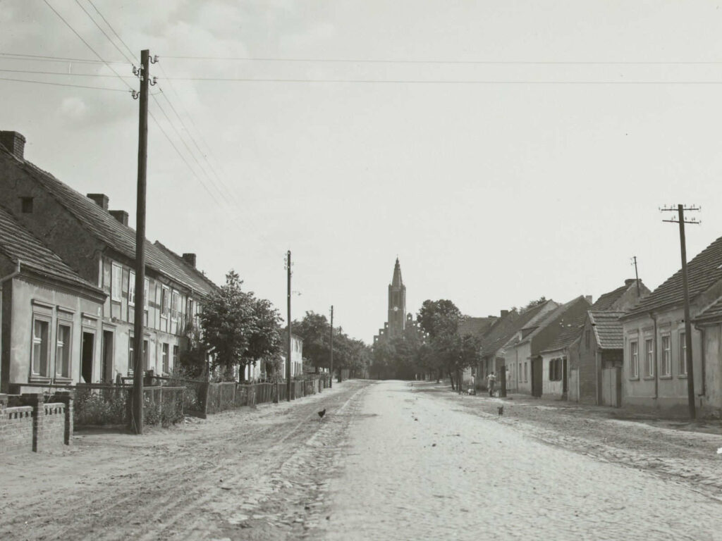 Bild aus 1934. Zeigt eine Straße. Links und rechts einstöckige Häuserreihe. Am Ende der Straße ragt ein Kirchenturm hervor. Vorne ist ein Strommast zu sehen.