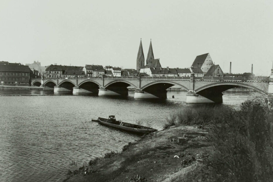 Das Foto ist vom Ufer der Oder aufgenommen. Im Mittelpunkt die lange Oderbrücke, im Vordergrund unbefestigter Boden, Sträucher und ein Boot, das im Wasser schwimmt. Im Hintergrund eine Stadt, aus der zwei Kirchtürme heraus ragen.