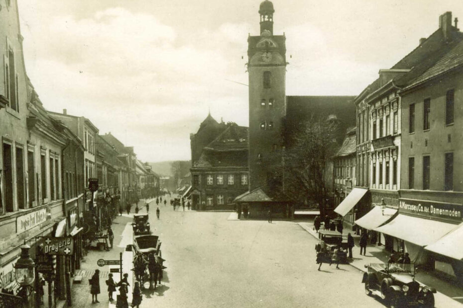 Schwarz-weiß Bild mit weißer Unterschrift "Fürstenwalde (Spree). Markt mit Rathaus). Zu sehen ist ein großer Platz, der in eine Straße über geht. Links und rechts sind mehrere Geschäfte abgebildet. Der Blick fällt auf das Rathaus, mit seinem herausragenden Turm und einer großen Uhr.