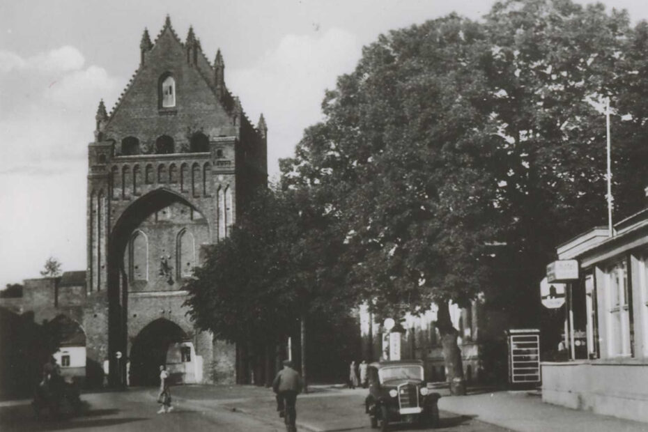 Zu sehen ist eine alte Postkarte, auf der das Ruppiner Tor und das Gesellschaftshaus abgebildet ist. In der Mitte eine große Straße, hinten im Bild ist das Tor mit mehreren Torbögen zu sehen.
