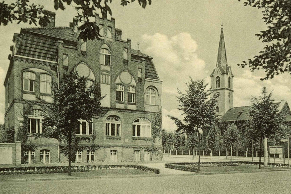Bild von 1942, auf dem sowohl das Rathaus als auch Kirche zu sehen sind. Im Vordergrund gepflasterte Straße, außerdem Rasenflächen, auf denen Bäume stehen und die von kleinen Hecken umzäunt werden.