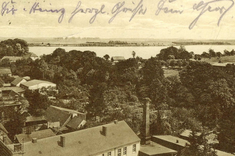 Postkarte mit Blick über Lindow auf den Wutzsee aus 1928. Zu sehen sind viele Dächer, Bäume und Wiesen.