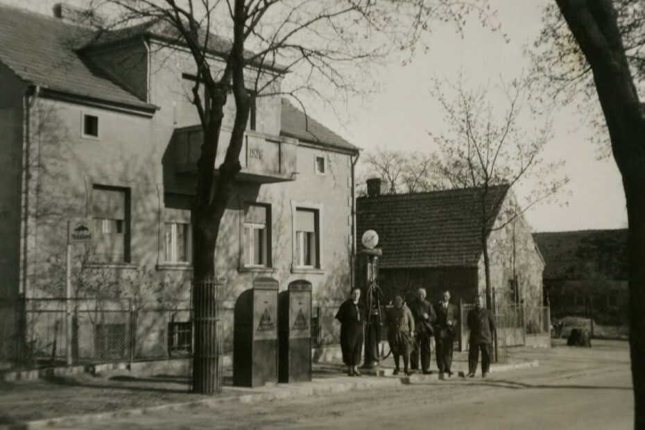Bild zeigt eine damalige Tankstelle mit einer sehr schmalen Tanksäule, um die mehrere Menschen herum stehen. Dahinter ein großes, alleinstehendes Wohnhaus.