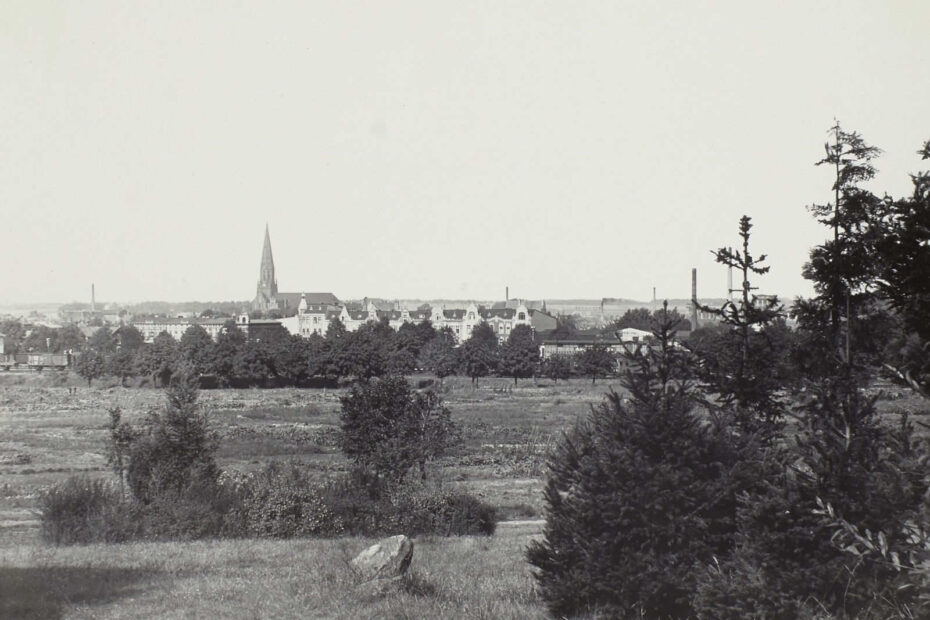 Bild von 1926, zeigt Pritzwalk aus etwas Entfernung von einer Wiese. Eine Stadt mit prächtigen, hohen Gebäuden und einem hochragenden Kirchtum ist zu sehen.