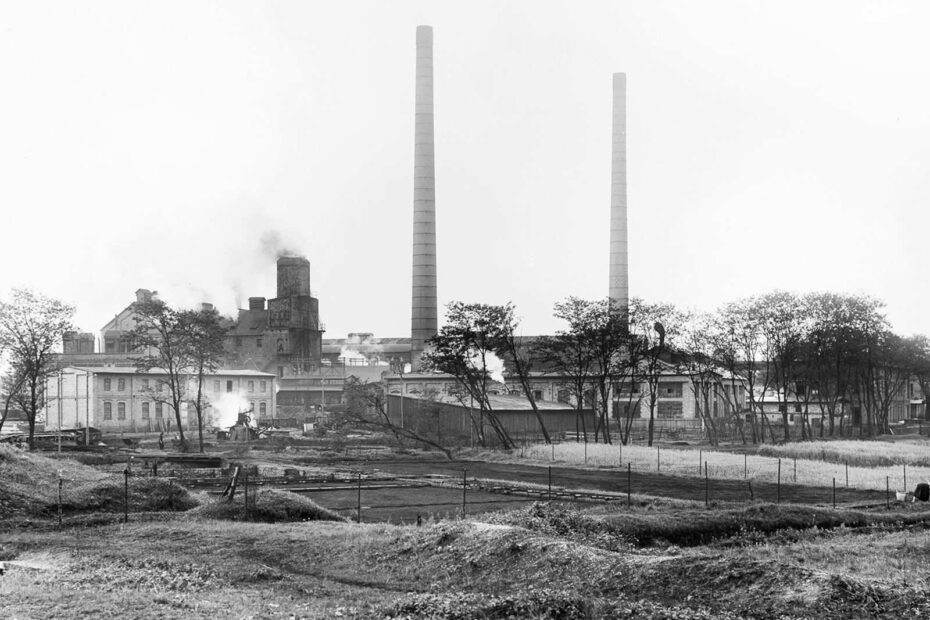 Bild von 1923, zeigt eine Fabrik aus der an verschiedenen Stellen Rauch raus steigt. Im Vordergrund Brachfläche, Zäune und kleine Erdhügel.