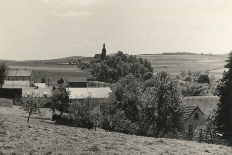 Bild wurde von einem Hügel aufgenommen. Es sind mehrere vereinzelte einfache Häuser zu sehen, eingerahmt von mehreren Feldern und Wiese. Weiter weg ein Kirchturm auf einer Erhebung zu sehen.