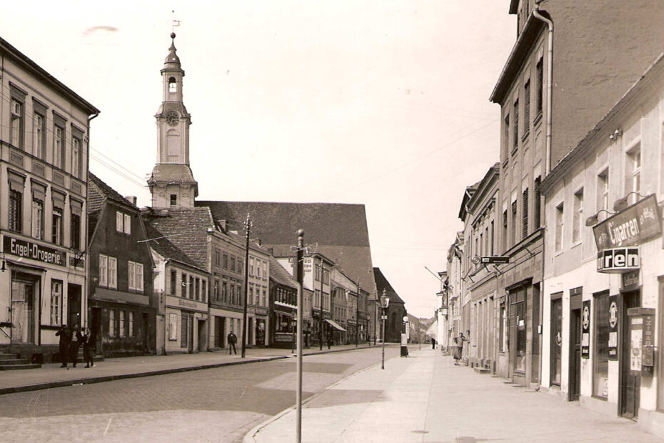 Bild aus der Ratsstraße in Wriezen. Eine Straße, rechts und links Häuser. An Geschäften sind eine Drogerie und ein Cigarren Geschäft zu erkennen.
